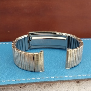 May include: A silver-toned metal expansion watch band featuring a textured, brushed finish on each rectangular link. The band includes a central folding clasp mechanism with a smooth, polished interior surface, designed for flexible wrist wear.