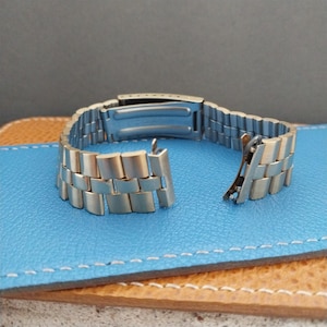 May include: A silver watch band with a clasp and a buckle. The band is made of metal links.