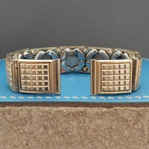 May include: A gold-tone metal watch band featuring a series of rectangular links with a raised grid pattern on the front-facing sections. The band has a flexible, expandable construction with silver-tone interior components visible between the links.