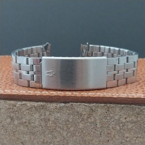 May include: A silver-toned watch band with a rectangular link design. The band features a brushed metal finish and a clasp with a logo. The watch band is displayed on a brown leather surface.