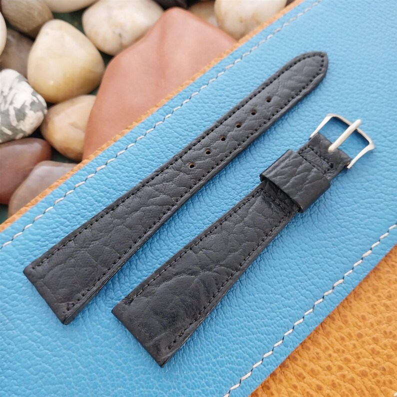 K&ouml;nnte beinhalten: Zwei schwarze Leder-Armb&auml;nder mit strukturierter Oberfl&auml;che. Eines der Armb&auml;nder hat eine silberfarbene Schnalle. Die Armb&auml;nder werden auf einer blau-beigen Oberfl&auml;che mit dekorativen N&auml;hten pr&auml;sentiert.