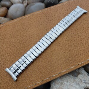 May include: A silver-colored, expandable watch band with a rectangular link design. The band is shown against a textured, brown leather surface. The watch band is made of metal and has a shiny finish. The band is designed to fit a watch.