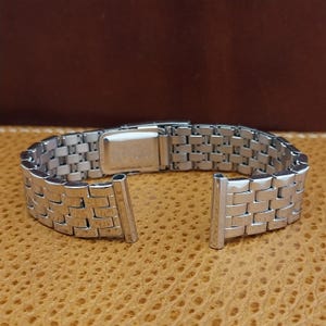 May include: A silver-toned metal watch band featuring a multi-link design with a polished finish. The bracelet includes a rectangular folding clasp mechanism and two straight end links, showcasing a classic construction with interconnected segments.