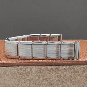 May include: A silver-toned watch band with a textured design. The band is composed of multiple rectangular links, each with a series of horizontal lines. The band is shown on a brown leather surface.