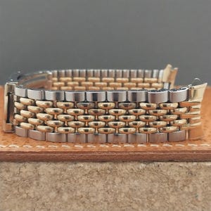 May include: A two-tone watch bracelet featuring a multi-link design with alternating silver-colored and gold-colored metal segments. The band includes a metallic clasp mechanism on one end and curved end links designed for attachment to a watch case.