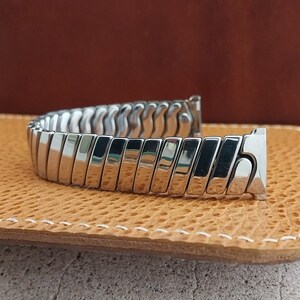May include: A silver-colored, expandable watch band. The band is made of rectangular, polished metal links that are connected in a flexible, accordion-like design. The band is curved and lies on a tan leather surface.