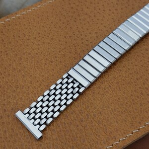 May include: A silver-colored watch band with a mesh design and rectangular links. The watch band is made of stainless steel and is displayed on a brown leather surface.