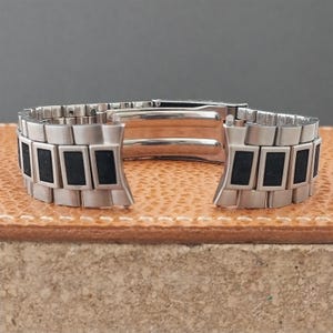 May include: A silver-toned metal watch bracelet featuring a multi-link design with rectangular black inlay accents on each link. The bracelet includes a central folding clasp mechanism with a smooth, polished interior finish and a secure locking system.