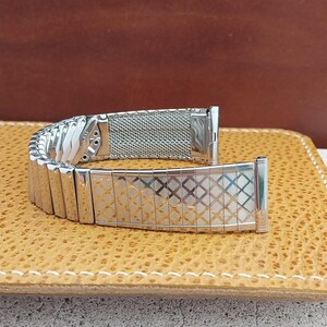 May include: A silver-tone metal watch band featuring a flexible expansion link design. The exterior surface displays a repeating diamond-shaped grid pattern with a polished finish, while the interior side shows a fine mesh texture.