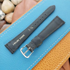 K&ouml;nnte beinhalten: Schwarzes Lederarmband mit silberner Schnalle. Auf dem Armband steht "ANILINE SHARK" und "R 3/4". Das Armband liegt auf einer blau-braunen Lederoberfl&auml;che.