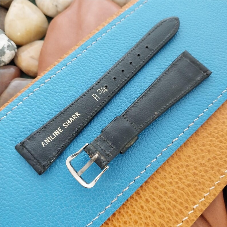 K&ouml;nnte beinhalten: Schwarzes Lederarmband mit silberner Schnalle. Auf dem Armband steht "ANILINE SHARK" und "R 3/4". Das Armband liegt auf einer blau-braunen Lederoberfl&auml;che.