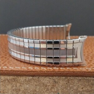 May include: A silver-toned, expandable watch band with a rectangular design. The band is composed of multiple small, rectangular links with a textured pattern. The band is resting on a brown surface.