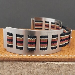 May include: A stainless steel watch band with a striped fabric strap in black, red, brown, and white. The band has the word "TISSOT" engraved on it. The watch band is curved and sits on a brown surface.