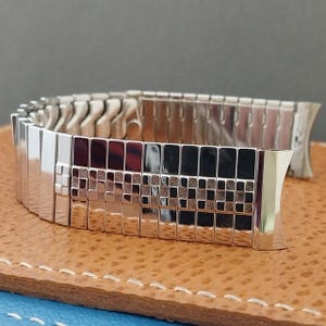 May include: A silver-toned metal expansion watch band featuring a central row of small, square, textured links flanked by smooth, polished rectangular links. The flexible, segmented design allows for easy adjustment and a comfortable fit on the wrist.