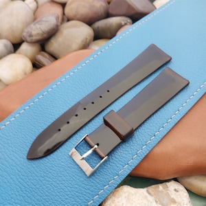 May include: A pair of dark brown watch straps with a glossy finish. The straps have a rectangular buckle made of silver-colored metal and are displayed on a blue leather surface. The straps are approximately 7 inches long.