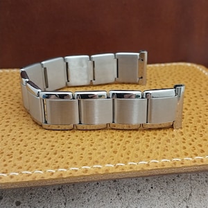 May include: A silver-toned metal watch band featuring a series of rectangular links with a brushed matte finish on the center panels and polished, reflective edges. The band is constructed with multiple articulated segments connected by pins.