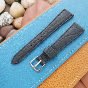 K&ouml;nnte beinhalten: Zwei schwarze Leder-Uhrenarmb&auml;nder mit strukturierter Oberfl&auml;che. Die Armb&auml;nder haben eine silberfarbene Schnalle und mehrere L&ouml;cher zur Gr&ouml;&szlig;enanpassung. Die Armb&auml;nder liegen auf einer blau-braunen Oberfl&auml;che.