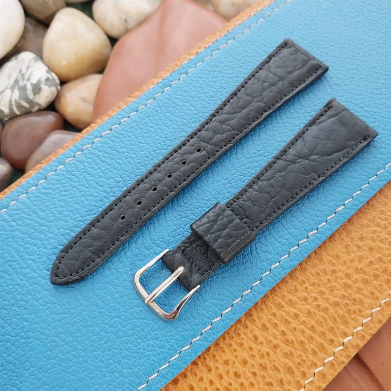 K&ouml;nnte beinhalten: Zwei schwarze Leder-Uhrenarmb&auml;nder mit strukturierter Oberfl&auml;che. Die Armb&auml;nder haben eine silberfarbene Schnalle und mehrere L&ouml;cher zur Gr&ouml;&szlig;enanpassung. Die Armb&auml;nder liegen auf einer blau-braunen Oberfl&auml;che.