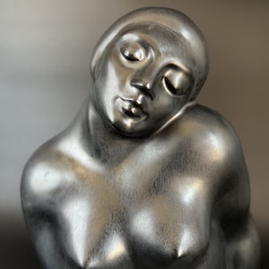 Può includere: Una scultura color argento di una figura reclinata con gli occhi chiusi. La scultura ha una finitura liscia e metallica e raffigura la parte superiore del corpo di una persona. La testa della figura è inclinata e il petto è leggermente esposto.