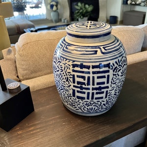 May include: A blue and white porcelain jar with a lid. The jar has a traditional Chinese design with a double happiness symbol on the lid. The jar is sitting on a dark brown wooden table.