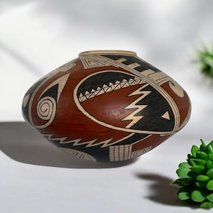 Puede incluir: Un jarrón de cerámica pintado a mano en rojo, negro y blanco con diseños geométricos. El jarrón es redondo y tiene una abertura ancha.