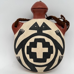 May include: A ceramic flask with a brown lid and neck, featuring a cream-colored body. The flask is adorned with black geometric designs, including figures and a cross-like shape. Brown rope handles are attached to the sides.