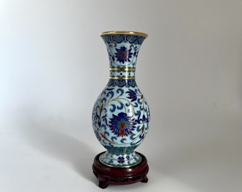 May include: A small blue and white cloisonné vase with a floral design. The vase is on a wooden stand.