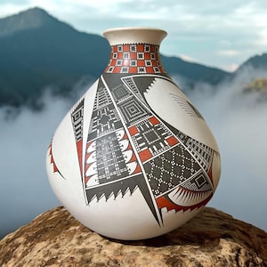 May include: A white ceramic vase with a geometric design in black and red. The vase has a rounded body and a narrow neck. The design features a variety of shapes and patterns, including triangles, squares, and diamonds.
