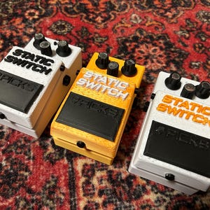 Puede incluir: Tres carcasas rectangulares para pedales de guitarra con perillas negras y un área de interruptor de pie negra. Dos son blancas con texto negro y naranja que dice STATIC SWITCH y PICKS, la central es amarilla con texto blanco.