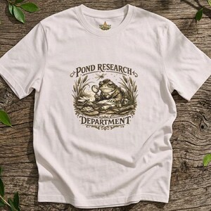 Op de afbeelding: Een wit t-shirt met korte mouwen en een bruine grafische print van een kikker met een hoed die een vergrootglas vasthoudt. De tekst POND RESEARCH DEPARTMENT staat in een decoratief lettertype rond de illustratie van de kikker.