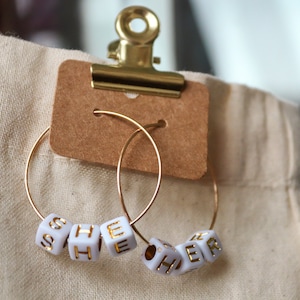 May include: A pair of gold hoop earrings with white beads spelling out "SHE" and "HER" in gold lettering.