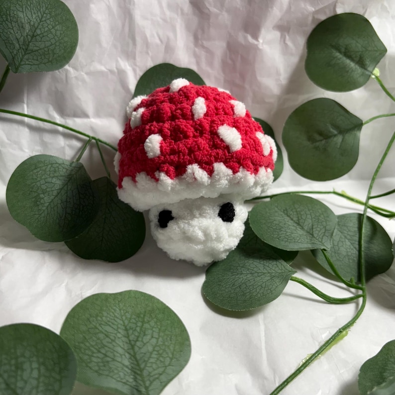 A handmade crochet mushroom toy with a red cap dotted with white spots. The stem is white, and it has two small black eyes. The mushroom is surrounded by green leaves.