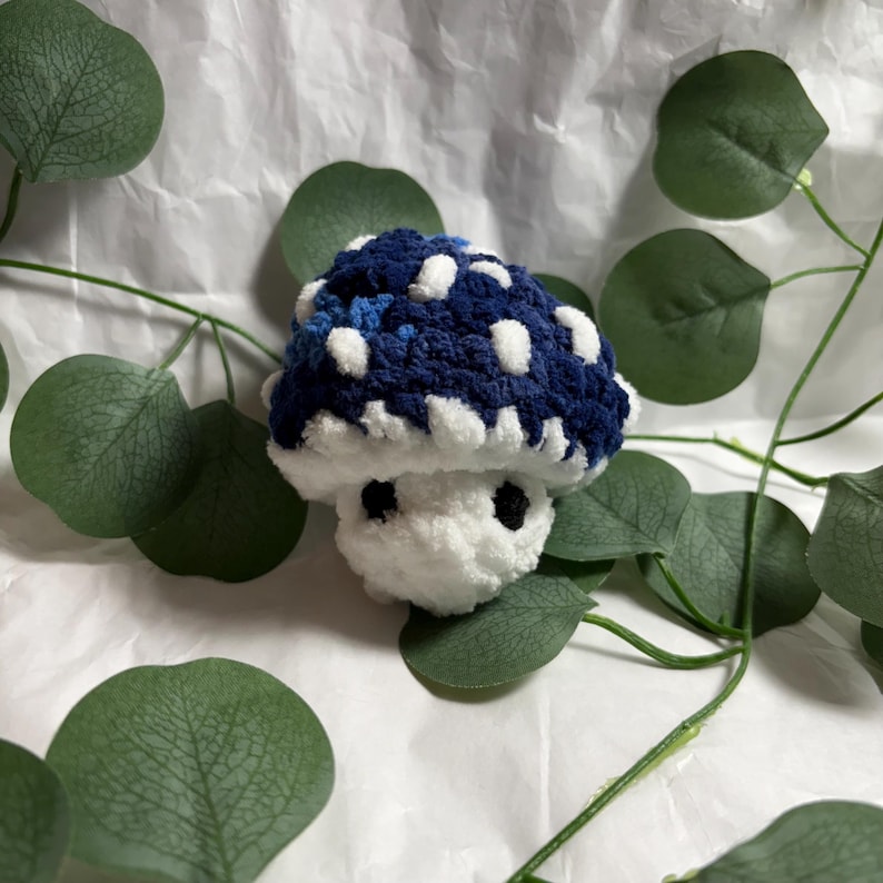 May include: A handmade crochet mushroom with a blue and white spotted cap and a white stem. The mushroom has two small black eyes and is surrounded by green leaves against a white background. This whimsical item is perfect for home decor.