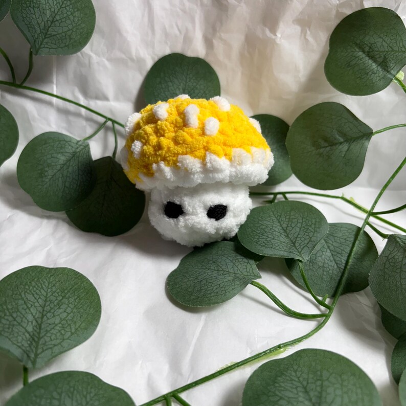 May include: A handmade, plush mushroom toy with a yellow cap dotted with white spots. The stem is white, and it has two black eyes. The toy is surrounded by green leaves and sits on a white surface.