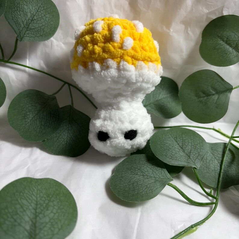 May include: A handmade crochet mushroom toy. The mushroom has a yellow cap with white spots, a white stem, and two black eyes. The toy is surrounded by green leaves on a white background.