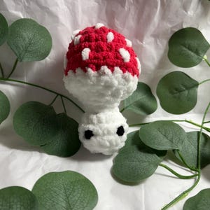 A handmade crochet mushroom toy with a red cap dotted with white spots. The stem is white, and it has two small black eyes. The mushroom is surrounded by green leaves.