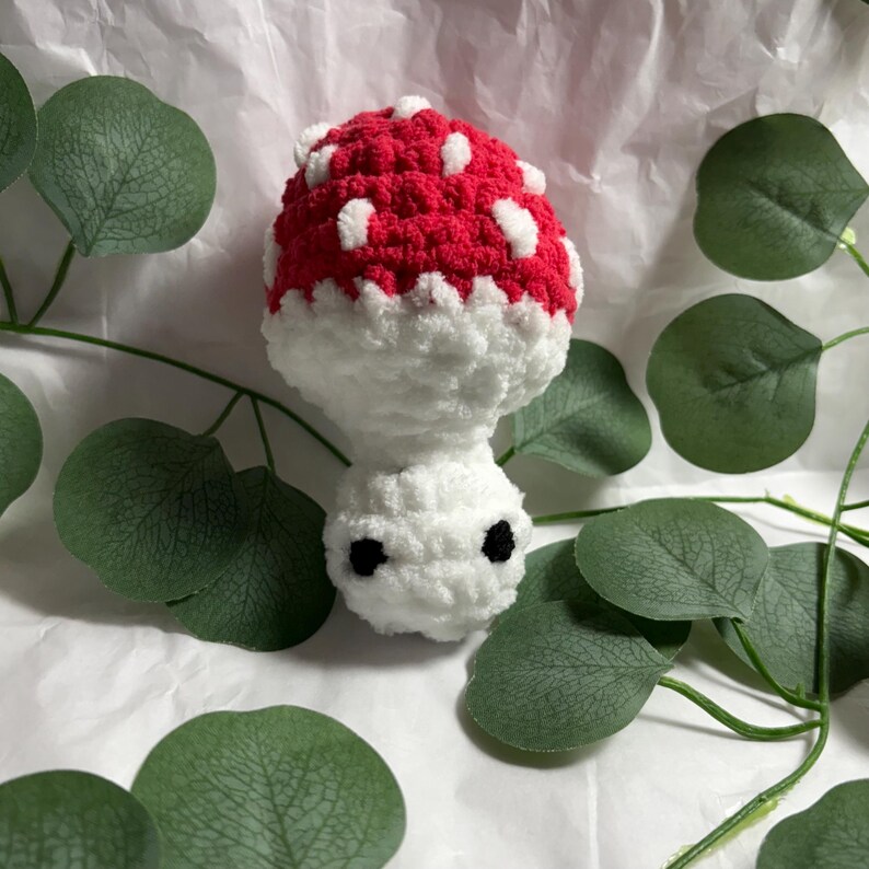 A handmade crochet mushroom toy with a red cap dotted with white spots. The stem is white, and it has two small black eyes. The mushroom is surrounded by green leaves.