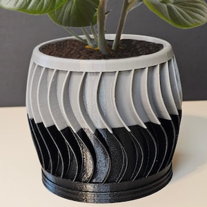 May include: A two-tone, 3D-printed planter with a unique design. The top half is light gray, transitioning to black at the bottom. The planter holds a green leafy plant with dark soil. The design features vertical, curved lines.