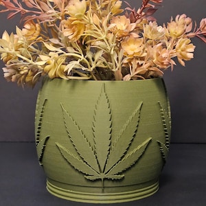 May include: A green, cylindrical planter with a cannabis leaf design. The planter holds a bouquet of faux flowers in shades of yellow, orange, and brown. The planter is made of a solid material and has a textured surface.