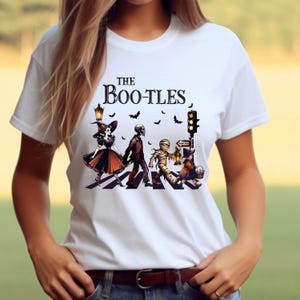 May include: White t-shirt with a graphic of a Halloween-themed parody of the Beatles' Abbey Road album cover. The graphic features a witch, a skeleton, a mummy, and a ghoul crossing a street.