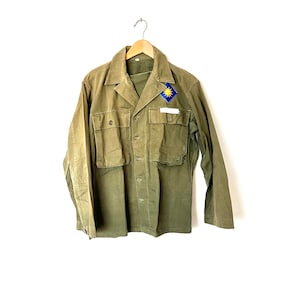 Può includere: Una giacca verde in stile militare con una toppa sul petto sinistro. La toppa presenta una stella gialla su sfondo blu.