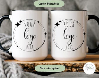 May include: Two white ceramic mugs with black handles and rims. Each mug features a circular graphic with four-pointed stars and the text "YOUR logo HERE" in the center. A green rectangular label above reads "Custom Photo/Logo" and one below reads "More color options".