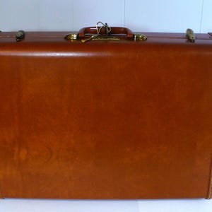 May include: A rectangular brown hard-shell suitcase featuring a top handle, gold-toned metal latches, and a central locking mechanism. The exterior has visible stitching along the edges, providing a structured appearance for this travel luggage.