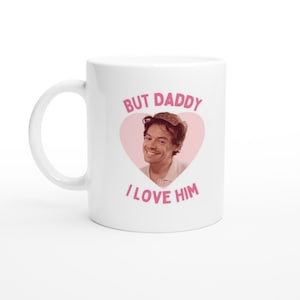Puede incluir: Taza de cerámica blanca con asa. La taza presenta un corazón rosa con una persona sonriente en su interior. El texto "BUT DADDY I LOVE HIM" está impreso en rosa encima y debajo del corazón.