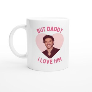 Puede incluir: Una taza de cerámica blanca con un gráfico de corazón rosa que contiene una fotografía de una persona sonriente con un blazer oscuro. El texto "BUT DADDY" está impreso en rosa sobre el corazón y "I LOVE HIM" en rosa debajo.