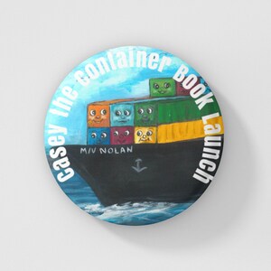 May include: A round button with a blue, white, and green illustration of a ship carrying colorful containers with faces. The text "Casey the Container Book Launch" is written around the edge of the button.