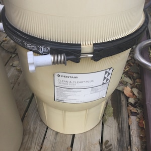 Pu&ograve; includere: Un filtro a cartuccia Pentair Clean & Clear Plus beige per piscine. Il filtro ha un coperchio nero e una maniglia bianca. Il filtro &egrave; posizionato su un ponte di legno.