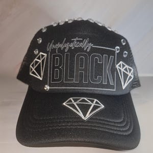 Op de afbeelding: Zwarte truckerpet met de tekst "Unapologetically BLACK" en diamantafbeeldingen. De pet is versierd met strass-steentjes langs de bovenrand. Hij heeft een mesh achterkant en een gebogen klep.