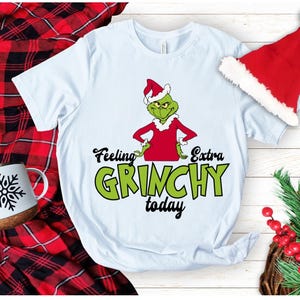 Puede incluir: Una camiseta de manga corta de color azul claro con un gráfico de un personaje verde que lleva un gorro y un abrigo de Papá Noel rojos. El texto dice Feeling Extra GRINCHY today en letras negras y verdes. La camiseta se muestra con un gorro de Papá Noel rojo.