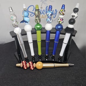Puede incluir: Siete bolígrafos coloridos con diferentes diseños en la parte superior de cada bolígrafo. Los bolígrafos se muestran en un soporte negro. El soporte está sobre una superficie negra. Hay un bolígrafo dorado con un diseño de tocino y huevo en la parte superior del bolígrafo en la parte delantera de la imagen.